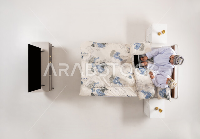 متابعة البرامج الترفيهية عبر اللابتوب، قضاء وقت ممتع مع الابناء، استخدام الأجهزة التقنية الحديثة، بورتريه علوي عامودي لرجل عربي خليجي عماني يرتدي الدشداشة والكمة يجلس على سرير مريح برفقة ابنه يحمل بيده الحاسوب المحمول، خلفيه بيضاء