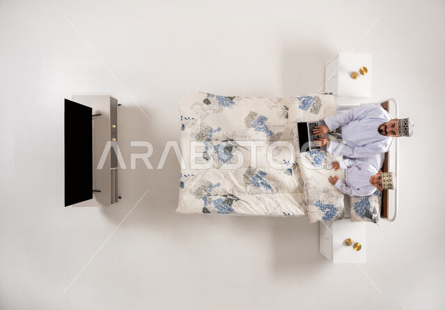 Spending quality time with children, using modern technology devices, watching entertainment programs on laptop, vertical overhead portrait of an Arab Gulf Omani man wearing a dishdasha and a kimono sitting on a comfortable bed with his son holding a laptop in his hand, white background