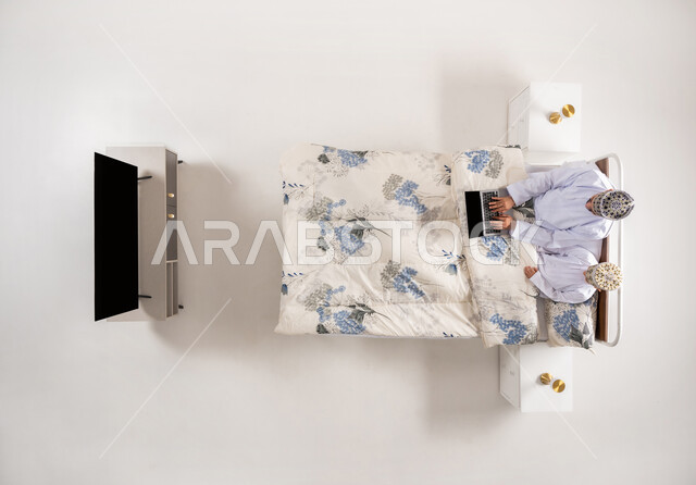 قضاء وقت ممتع مع الابناء، استخدام الأجهزة التقنية الحديثة، متابعة البرامج الترفيهية عبر اللابتوب، بورتريه علوي عامودي لرجل عربي خليجي عماني يرتدي الدشداشة والكمة يجلس على سرير مريح برفقة ابنه يحمل بيده الحاسوب المحمول، خلفيه بيضاء