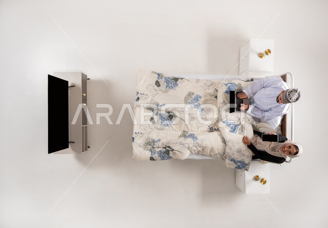 إنجاز مهام الأعمال عن بعد، استخدام الأجهزة التقنية المتطورة، دمج التكنولوجيا الحديثة بالحياة العملية، بورتريه علوي عامودي لرجل عربي خليجي عماني يرتدي الدشداشة والكمة يعمل على جهاز الحاسوب المحمول يجلس على سرير مريح بجانب زوجته، امرأة تحمل بيدها الجوال، خلفيه بيضاء