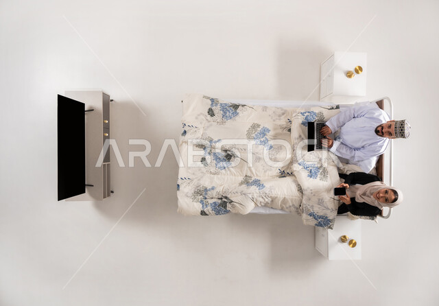 Completing business tasks remotely, using advanced technical devices, integrating modern technology into work life, vertical overhead portrait of an Arab Gulf Omani man wearing a dishdasha and a turban working on a laptop sitting on a comfortable bed next to his wife, a woman holding a mobile phone in her hand, white background