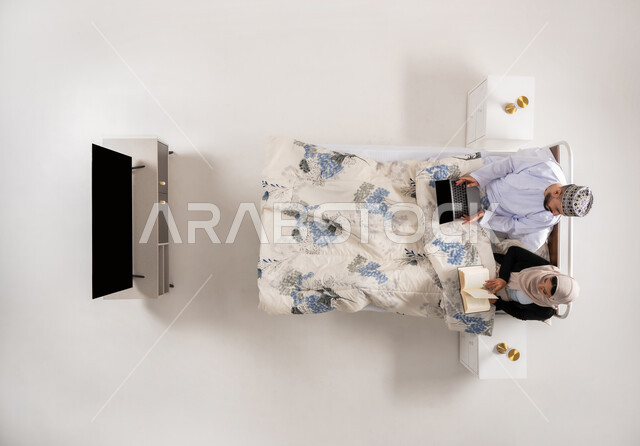 استخدام الأجهزة التقنية الحديثة، مفهوم التثقيف الذاتي وحب المطالعة، انجاز المهام عن بعد، بورتريه علوي عامودي لرجل عربي خليجي عماني يرتدي الدشداشة والكمة يعمل على جهاز الحاسوب يجلس على سرير مريح برفقة زوجته، امرأة تمسك كتاب تقوم بممارسة هواية القراءة، خلفية بيضاء