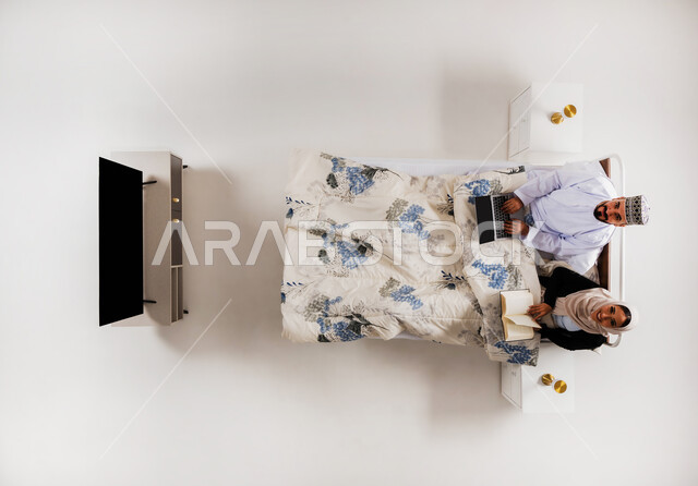 استخدام الأجهزة التقنية الحديثة، مفهوم التثقيف الذاتي وحب المطالعة، انجاز المهام عن بعد، بورتريه علوي عامودي لرجل عربي خليجي عماني يرتدي الدشداشة والكمة يعمل على جهاز الحاسوب يجلس على سرير مريح برفقة زوجته، امرأة تمسك كتاب تقوم بممارسة هواية القراءة، خلفية بيضاء