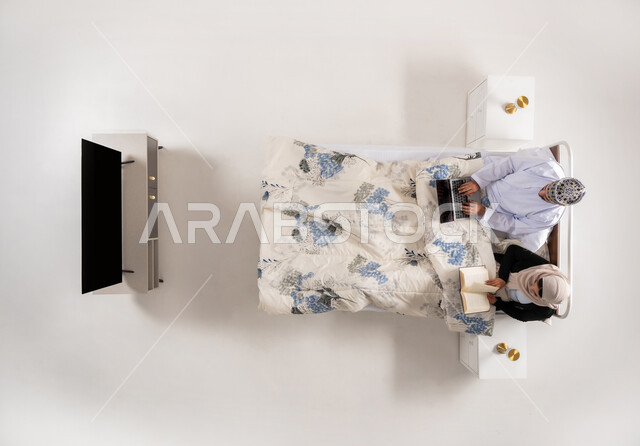 مفهوم التثقيف الذاتي وحب المطالعة، انجاز المهام عن بعد، استخدام الأجهزة التقنية الحديثة، بورتريه علوي عامودي لرجل عربي خليجي عماني يرتدي الدشداشة والكمة يعمل على جهاز الحاسوب يجلس على سرير مريح برفقة زوجته، امرأة تمسك كتاب تقوم بممارسة هواية القراءة، خلفية بيضاء
