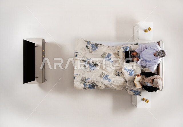 انجاز المهام عن بعد، مفهوم التثقيف الذاتي وحب المطالعة، استخدام الأجهزة التقنية الحديثة، بورتريه علوي عامودي لرجل عربي خليجي عماني يرتدي الدشداشة والكمة يعمل على جهاز الحاسوب يجلس على سرير مريح برفقة زوجته، امرأة تمسك كتاب تقوم بممارسة هواية القراءة، خلفية بيضاء