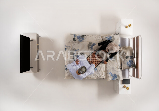 قضاء وقت ممتع مع العائلة، مفهوم التركيز والتحدي, مفهوم التخطيط والتفكير، بورتريه عامودي علوي لزوجين عربيين خليجيين عمانيين يجلسان على سرير مريح يلعبان لعبة الشطرنج، ألعاب ثنائية استراتيجية، خلفية بيضاء