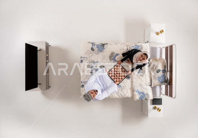 Concept of planning and thinking, looking at the camera with expressions of pleasure and happiness, having a good time with family, concept of focus and challenge, vertical top portrait of an Arab Gulf Omani couple sitting on a comfortable bed playing chess, strategic dual games, white background