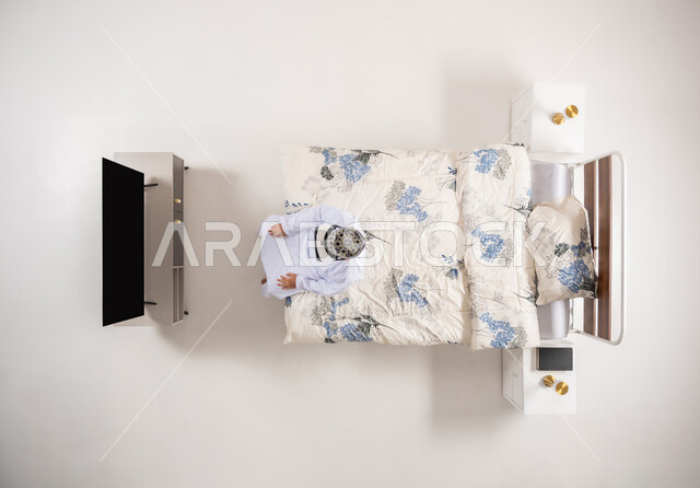 قضاء أوقات ممتعة ومسلية بالألعاب الافتراضية، الاستمتاع بمشاهدة أفلام الواقع الافتراضي، الاندماج ومحاكاة العالم الخيالي، بورتريه علوي عامودي لرجل عربي خليجي عماني يجلس على سرير مريح يرتدي الدشداشة والكمة ونظارات (الفي ار) ثلاثية الأبعاد، خلفية بيضاء