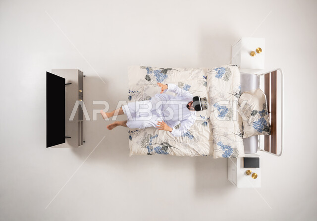 قضاء أوقات ممتعة ومسلية بالألعاب الافتراضية، الاستمتاع بمشاهدة أفلام الواقع الافتراضي، الاندماج ومحاكاة العالم الخيالي، بورتريه علوي عامودي لرجل عربي خليجي عماني يجلس على سرير مريح يرتدي الدشداشة والكمة ونظارات (الفي ار) ثلاثية الأبعاد، خلفية بيضاء