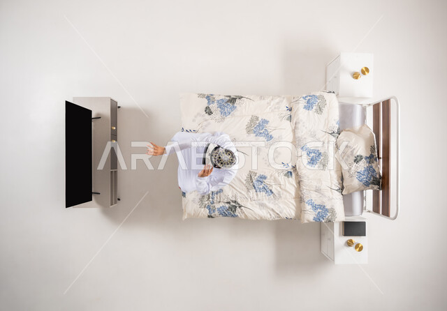 محاكاة العالم الخيالي، قضاء أوقات ممتعة ومسلية بالألعاب الافتراضية، الاستمتاع بمشاهدة أفلام الواقع الافتراضي، بورتريه علوي عامودي لرجل عربي خليجي عماني يجلس على سرير مريح يرتدي الدشداشة والكمة ونظارات (الفي ار) ثلاثية الأبعاد، خلفية بيضاء