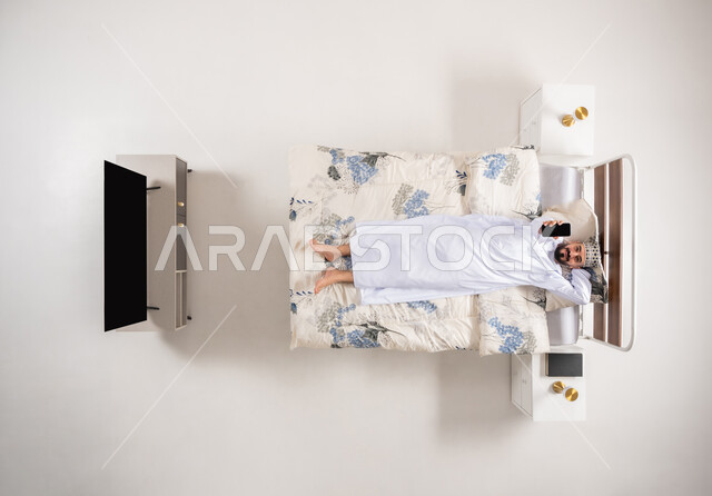 استخدام الاجهزة التقنية الحديثة، الاستفادة من خدمات وتطبيقات الجوال، بورتريه علوي عامودي لرجل عربي خليجي عماني يجلس على سرير مريح يحمل بيده الهاتف المحمول يقوم بعرض شاشة سوداء فارغة، خلفية بيضاء