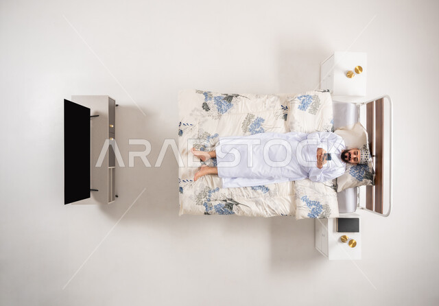 Chatting with family and friends, browsing social networking sites, taking advantage of mobile services and applications, vertical top portrait of an Arab Gulf Omani man sitting on a comfortable bed holding a mobile phone in his hand, online marketing, white background