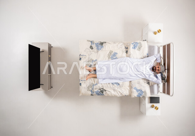 الشعور بالتعب والخمول والحاجة للنوم, إيماءات الراحة والإسترخاء، بورتريه علوي عامودي لرجل عربي خليجي عماني يرتدي الدشداشة والكمة يضع يديه اسفل رأسه ويستلقي على سرير مريح، تعابير التأمل والتفكير، خلفية بيضاء