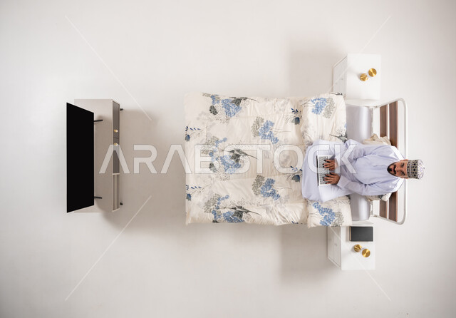 استخدام الأجهزة التقنية المتطورة، متابعة المشاريع والخطط عبر الانترنت، إنجاز مهام الأعمال عن بعد، دمج التكنولوجيا الحديثة بالحياة العملية، بورتريه علوي عامودي لرجل عربي خليجي عماني يرتدي الدشداشة والكمة يجلس على سرير مريح يعمل على جهاز الحاسوب المحمول، خلفيه بيضاء