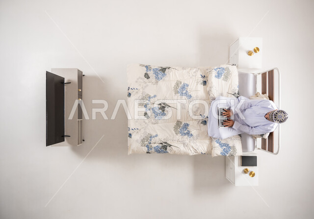 متابعة المشاريع والخطط عبر الانترنت، إنجاز مهام الأعمال عن بعد، استخدام الأجهزة التقنية المتطورة، دمج التكنولوجيا الحديثة بالحياة العملية، بورتريه علوي عامودي لرجل عربي خليجي عماني يرتدي الدشداشة والكمة يجلس على سرير مريح يعمل على جهاز الحاسوب المحمول، خلفيه بيضاء