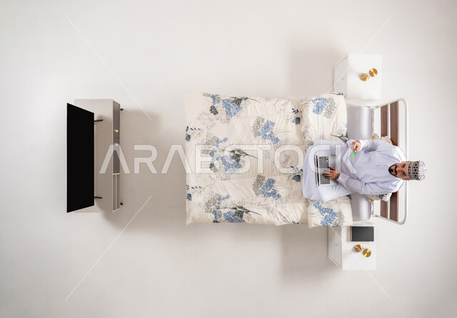 الدفع عبر تطبيقات اللابتوب، إمكانية تسديد الفواتير عبر البرامج الالكترونية، استخدام البطاقة البنكية الالكترونية، بورتريه علوي عامودي لرجل عربي خليجي عماني يرتدي الدشداشة والكمة يجلس على سرير مريح يحمل بيده جهاز الحاسوب المحمول، خلفية بيضاء