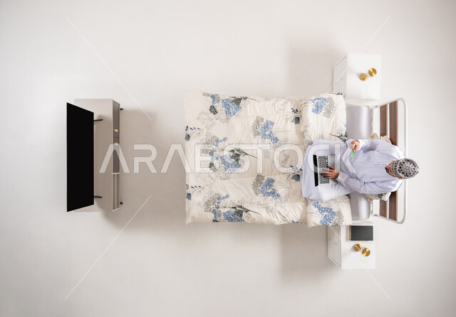 استخدام البطاقة البنكية الالكترونية، إمكانية تسديد الفواتير عبر البرامج الالكترونية، الدفع عبر تطبيقات اللابتوب، بورتريه علوي عامودي لرجل عربي خليجي عماني يرتدي الدشداشة والكمة يجلس على سرير مريح يحمل بيده جهاز الحاسوب المحمول، خلفية بيضاء