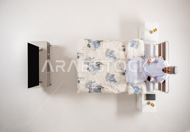 الدفع عبر تطبيقات الجوال، إمكانية تسديد الفواتير عبر البرامج الالكترونية، استخدام البطاقة البنكية الالكترونية، بورتريه علوي عامودي لرجل عربي خليجي عماني يرتدي الدشداشة والكمة يجلس على سرير مريح يحمل بيده الهاتف المحمول، خلفية بيضاء