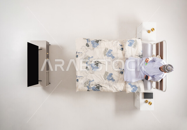 استخدام البطاقة البنكية الالكترونية، الدفع عبر تطبيقات الجوال، إمكانية تسديد الفواتير عبر البرامج الالكترونية، بورتريه علوي عامودي لرجل عربي خليجي عماني يرتدي الدشداشة والكمة يجلس على سرير مريح يحمل بيده الهاتف المحمول، خلفية بيضاء