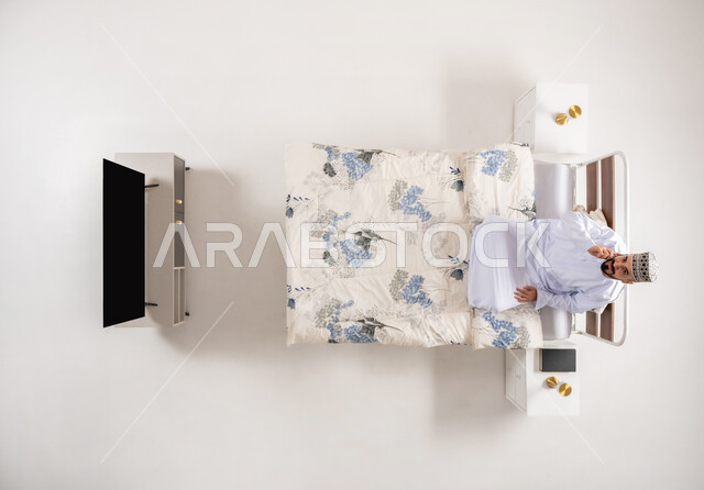 استخدام الاجهزة التقنية المتطورة، إجراء مكالمة هاتفية مع الأهل والأصدقاء، بورتريه علوي عامودي لرجل عربي خليجي عماني يرتدي الدشداشة والكمة يجلس على سرير مريح يحمل بيده الهاتف المحمول، دمج التكنولوجيا الحديثة بالحياة اليومية، خلفية بيضاء