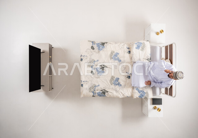 دمج التكنولوجيا الحديثة بالحياة اليومية، استخدام الاجهزة التقنية المتطورة، إجراء مكالمة هاتفية مع الأهل والأصدقاء، بورتريه علوي عامودي لرجل عربي خليجي عماني يرتدي الدشداشة والكمة يجلس على سرير مريح يحمل بيده الهاتف المحمول، خلفية بيضاء