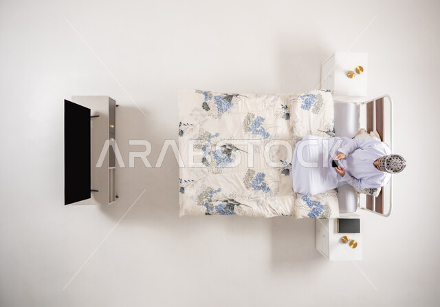 استخدام الأجهزة والتقنيات المتطورة، التواصل مع الأهل والأصدقاء، التسوق عبر الانترنت، تصفح برامج و تطبيقات الجوال، بورتريه  علوي عامودي لرجل عربي خليجي عماني يرتدي الدشداشة والكمة يجلس على سرير مريح يحمل بيده الهاتف المحمول، خلفية بيضاء