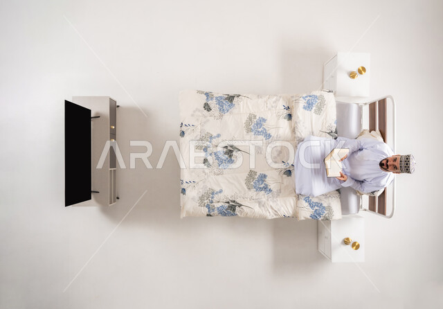 قضاء وقت الفراغ بالتعلم والثقافة، مفهوم التثقيف والبحث العلمي، بورتريه علوي عامودي لرجل عربي خليجي عماني يرتدي الدشداشة والكمة يجلس على سرير مريح يقوم قراءة كتاب, هواية المطالعة والاستكشاف، خلفية بيضاء