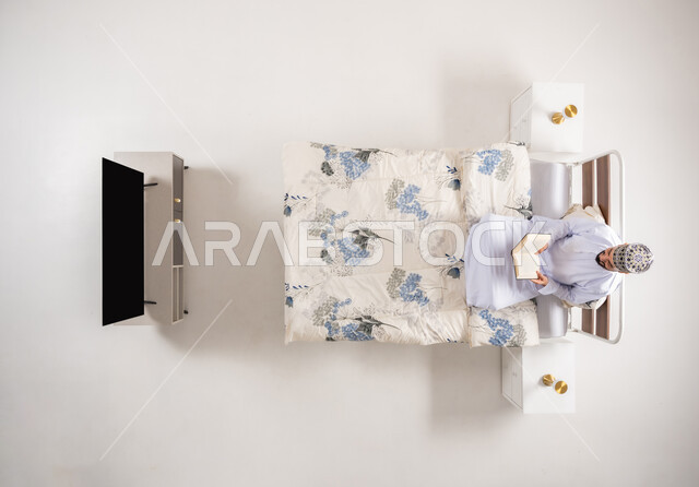 هواية المطالعة والاستكشاف، مفهوم التثقيف والبحث العلمي، قضاء وقت الفراغ بالتعلم والثقافة، بورتريه علوي عامودي لرجل عربي خليجي عماني يرتدي الدشداشة والكمة يجلس على سرير مريح يقوم قراءة كتاب, خلفية بيضاء