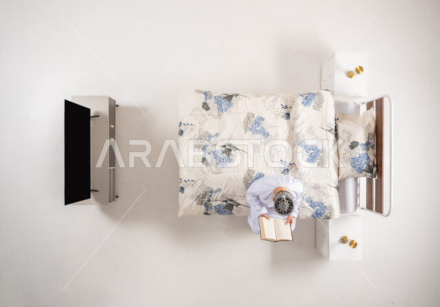 قضاء وقت الفراغ بالتعلم والثقافة، مفهوم التثقيف والبحث العلمي، بورتريه علوي عامودي لرجل عربي خليجي عماني يرتدي الدشداشة والكمة يجلس على سرير مريح يقوم قراءة كتاب, هواية المطالعة والاستكشاف، خلفية بيضاء