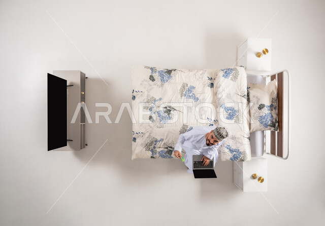 إمكانية تسديد الفواتير عبر البرامج الالكترونية، استخدام البطاقة البنكية الالكترونية، الدفع عبر تطبيقات اللابتوب، بورتريه علوي عامودي لرجل عربي خليجي عماني يرتدي الدشداشة والكمة يجلس على سرير مريح يحمل بيده جهاز الحاسوب المحمول، خلفية بيضاء