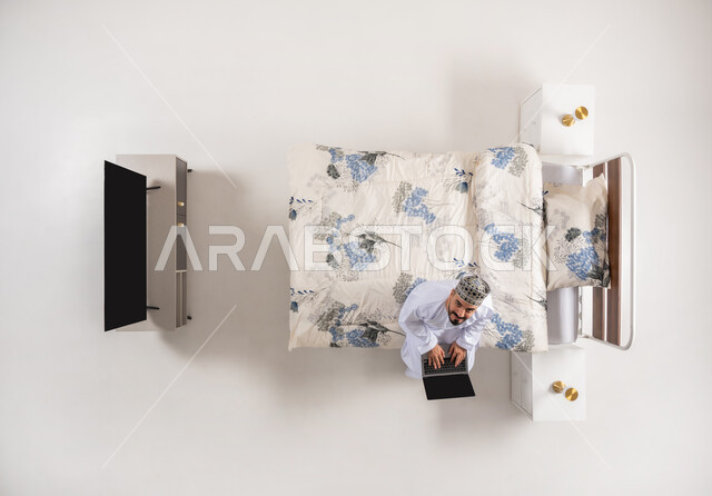 متابعة المشاريع والخطط عبر الانترنت، إنجاز مهام الأعمال عن بعد، استخدام الأجهزة التقنية المتطورة، دمج التكنولوجيا الحديثة بالحياة العملية، بورتريه علوي عامودي لرجل عربي خليجي عماني يرتدي الدشداشة والكمة يجلس على سرير مريح يعمل على جهاز الحاسوب المحمول، خلفيه بيضاء