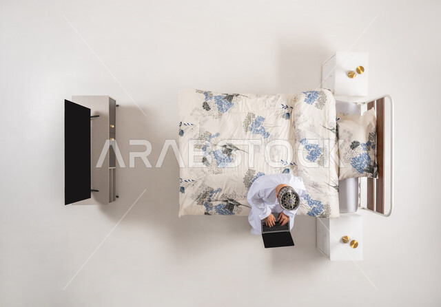 إنجاز مهام الأعمال عن بعد، متابعة المشاريع والخطط عبر الانترنت، استخدام الأجهزة التقنية المتطورة، دمج التكنولوجيا الحديثة بالحياة العملية، بورتريه علوي عامودي لرجل عربي خليجي عماني يرتدي الدشداشة والكمة يجلس على سرير مريح يعمل على جهاز الحاسوب المحمول، خلفيه بيضاء