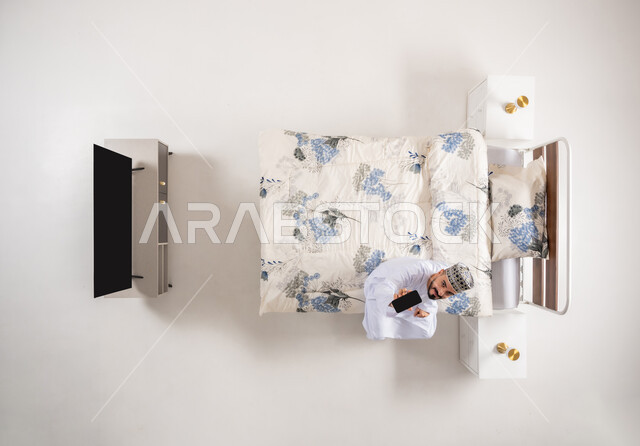 Showing a blank black screen, using advanced devices and technologies, taking advantage of mobile programs and applications, vertical overhead portrait of an Arab Gulf Omani man wearing a dishdasha and a kimono sitting on a comfortable bed holding a mobile phone in his hand, white background