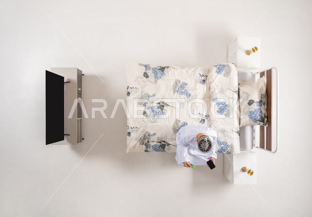 Online shopping, communicating with family and friends, browsing mobile programs and applications, vertical overhead portrait of an Arab Gulf Omani man wearing a dishdasha and a kummah sitting on a comfortable bed holding a mobile phone in his hand, using advanced devices and technologies, white background