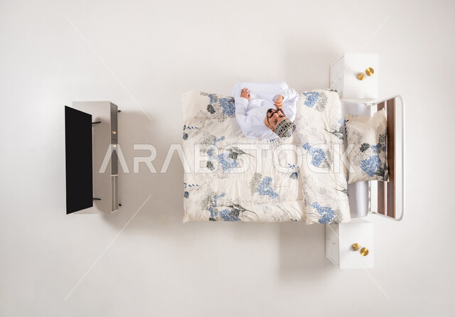 استخدام الاجهزة التقنية المتطورة، إجراء مكالمة هاتفية مع الأهل والأصدقاء، بورتريه علوي عامودي لرجل عربي خليجي عماني يرتدي الدشداشة والكمة يجلس على سرير مريح يحمل بيده الهاتف المحمول، دمج التكنولوجيا الحديثة بالحياة اليومية، خلفية بيضاء