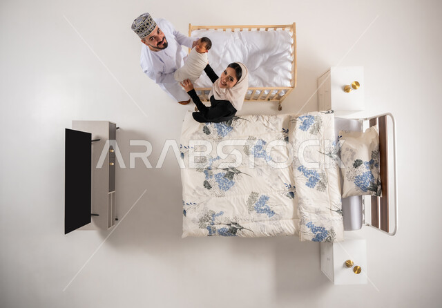 مداعبه الأبناء بحب وحنان، قضاء الوقت باللعب مع الاولاد، مفهوم الاسرة وتحمل المسؤولية، العناية بالاطفال حديثي الولاده، بورتريه علوي عامودي لاب وام عربيان خليجيان عمانيان يحملان طفلهما الرضيع بحرص واهتمام، خلفية بيضاء