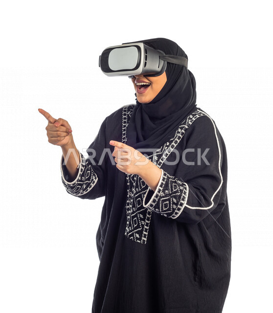 بورتريه مرأة عربية سعودية خليجية ترتدي نظارة الواقع الإفتراضي VR ثلاثية الأبعاد الفريدة ، تشير بيدها بأصبع السبابة لشيئ ما تشاهده ، التفاعل و المتعة و التسلية بمشاهدة الأفلام و ألعاب العالم الافتراضي ، تقنيات و ألعاب ، خلفية بيضاء