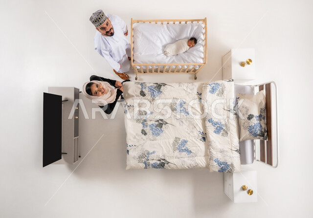 مفهوم الاسرة وتحمل المسؤولية، العناية بالاطفال حديثي الولاده، مداعبه الأبناء بحب وحنان، قضاء الوقت باللعب مع الاولاد، بورتريه علوي عامودي لاب وام عربيان خليجيان عمانيان يقفان بجانب سرير طفلهما الرضيع ينظران الى الكاميرا بايماءات السرور والسعادة، خلفية بيضاء
