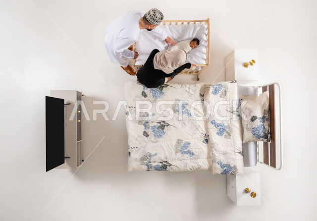 العناية بالاطفال حديثي الولاده، مداعبه الأبناء بحب وحنان، قضاء الوقت باللعب مع الاولاد، مفهوم الاسرة وتحمل المسؤولية، بورتريه علوي عامودي لاب وام عربيان خليجيان عمانيان يضعان طفلهما الرضيع بالسرير بحرص واهتمام، خلفية بيضاء