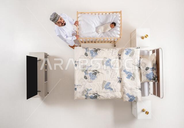 مفهوم الاسرة وتحمل المسؤولية، تقديم مشاعر الحب والحنان للاولاد، العناية بالاطفال حديثي الولاده، دور الأهل في حياه الأبناء، الرعاية والاهتمام بالأبناء، بورتريه علوي عامودي لاب عربي خليجي عماني يرتدي الدشداشة والكمة يقوم بهزسرير طفله الرضيع، خلفيه بيضاء