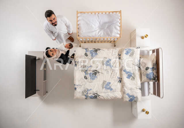 ايماءات السرور والسعادة، مفهوم الاسرة وتحمل المسؤولية، العناية بالاطفال حديثي الولاده، مداعبه الأبناء بحب وحنان، قضاء الوقت باللعب مع الاولاد، بورتريه علوي عامودي لام عربية خليجية عمانية تحمل بيدها طفلها تقف بجانب زوجها، عائلة سعودية تقوم برعاية ابنها، خلفية بيضاء