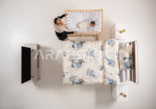 النظر للكاميرا بتعابير السرور والسعادة، العناية بالاطفال حديثي الولاده، دور الأهل في حياه الأبناء، امرأة سعودية تقوم بملاطفة ولدها، بورتريه علوي عامودي لام عربيه خليجيه عمانية بهز مهد طفلها، تقديم مشاعر الحب والحنان للاولاد، مفهوم الاسرة وتحمل المسؤولية، خلفيه بيضاء