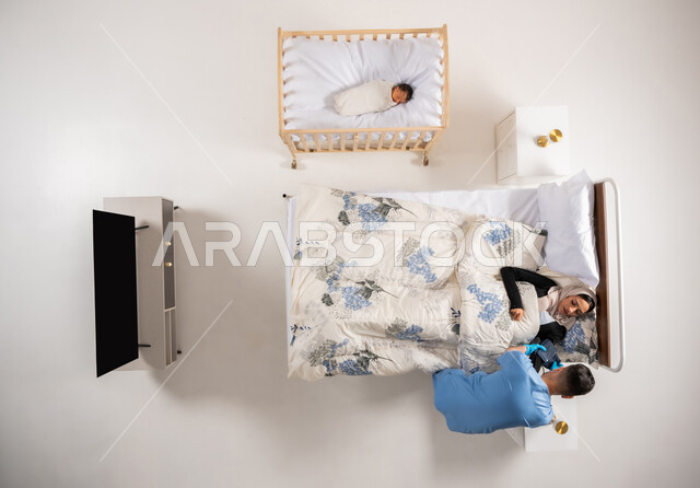 تلقي الرعايه الطبيه في المنزل، اجراء فحوصات ما بعد الولادة، بورتريه علوي عامودي لممرض عربي خليجي سعودي يقوم بفحص الضغط(جهاز السفينجومانوميتر) لسيده مريضه مستلقيه على سريرها، مفهوم العلاج والعناية الصحية، شاب عماني يعمل في مجال الطب، خلفيه بيضاء