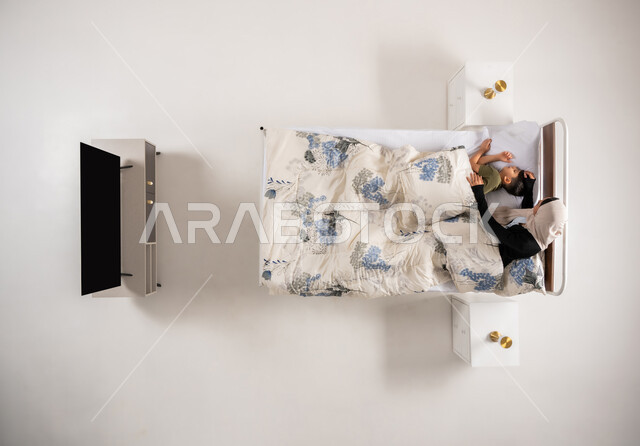 مفهوم العناية والاهتمام بالأولاد، بورتريه علوي عامودي لأم عربية خليجية عمانية مستلقية على سرير تقوم بمداعبة ابنها، امرأة سعودي محجبة ترعى ولدها، فوائد النوم المبكر على صحة الصغار، الاسترخاء والاستمتاع بالأجواء الهادئة، اتباع أسلوب حياة صحي، خلفية بيضاء