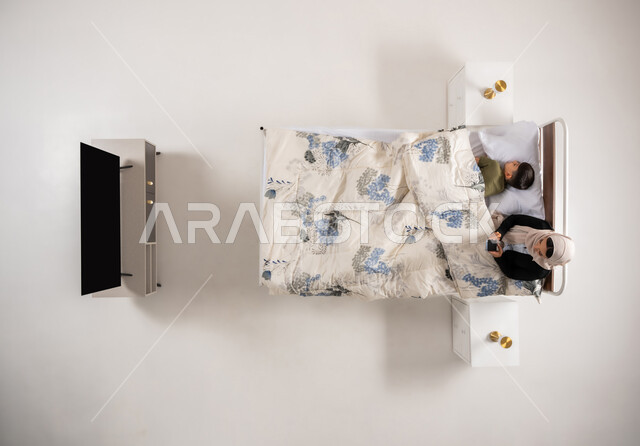 الدردشة والتواصل مع الاهل والاصدقاء، اهمية النوم المبكر، بورتريه علوي عامودي لأم عربية خليجية عمانية مستلقية على سرير مريح بجانب ابنها تحمل بيدها الهاتف المحمول تتصفح مواقع التواصل الاجتماعي، امرأة سعودي محجبة تقوم باستخدام التقنيات الحديثة، خلفية بيضاء