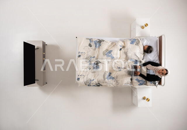 Accomplishing tasks and work, using modern and advanced technological devices, vertical upper portrait of a veiled Gulf Arab Omani woman sitting on a comfortable bed working on a laptop, a young Saudi woman following up on projects and plans online, white background
