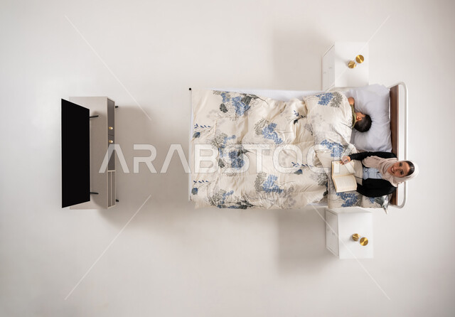 قراءه القصص المفيده للأبناء قبل النوم، مفهوم التربيه القائمه على الحب والحنان، بورتريه علوي عامودي لامرأة عربية خليجية عمانية تقرأ الروايات المسليه لابنها، سيدة سعودية محجبة تستلقي على سرير مريح برفقة ولدها، قضاء الوقت مع الابن، خلفية بيضاء