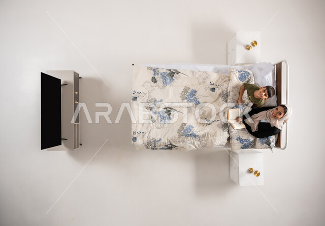 مفهوم التربيه القائمه على الحب والحنان، قراءه القصص المفيده للأبناء قبل النوم، بورتريه علوي عامودي لامرأة عربية خليجية عمانية تقرأ الروايات المسليه لابنها، سيدة سعودية محجبة تستلقي على سرير مريح برفقة ولدها، قضاء الوقت مع الابن، خلفية بيضاء