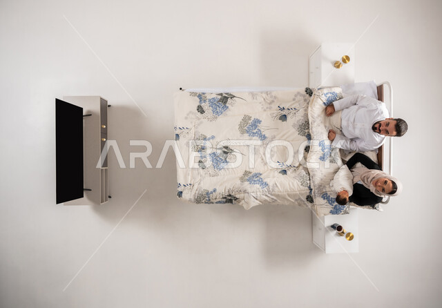 النظر الى الكاميرا بإيماءات السرور، تعزيز الروابط الاسرية، أجواء عائلية مليئة بالحب والحنان، سيدة عمانية تحمل رضيعها حديث الولادة بإيماءات العطف، بورتريه عامودي علوي لإمرأه ورجل عربيان سعوديان خليجيان يجلسان على سرير مريح ينظران الى طفلهما الصغير، خلفية بيضاء