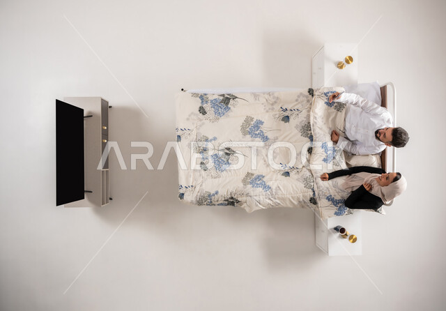 الشعور بالفرح والسعادة، توثيق اللحظات السعيدة، رجل عماني يمسك بيده الهاتف المحمول يقوم بالتقاط صور السيلفي التذكارية، بورتريه عامودي علوي لإمرأه وشاب عربيان سعوديان خليجيان يجلسان على سرير مريح يستخدمان الأجهزة التقنية، خلفية بيضاء